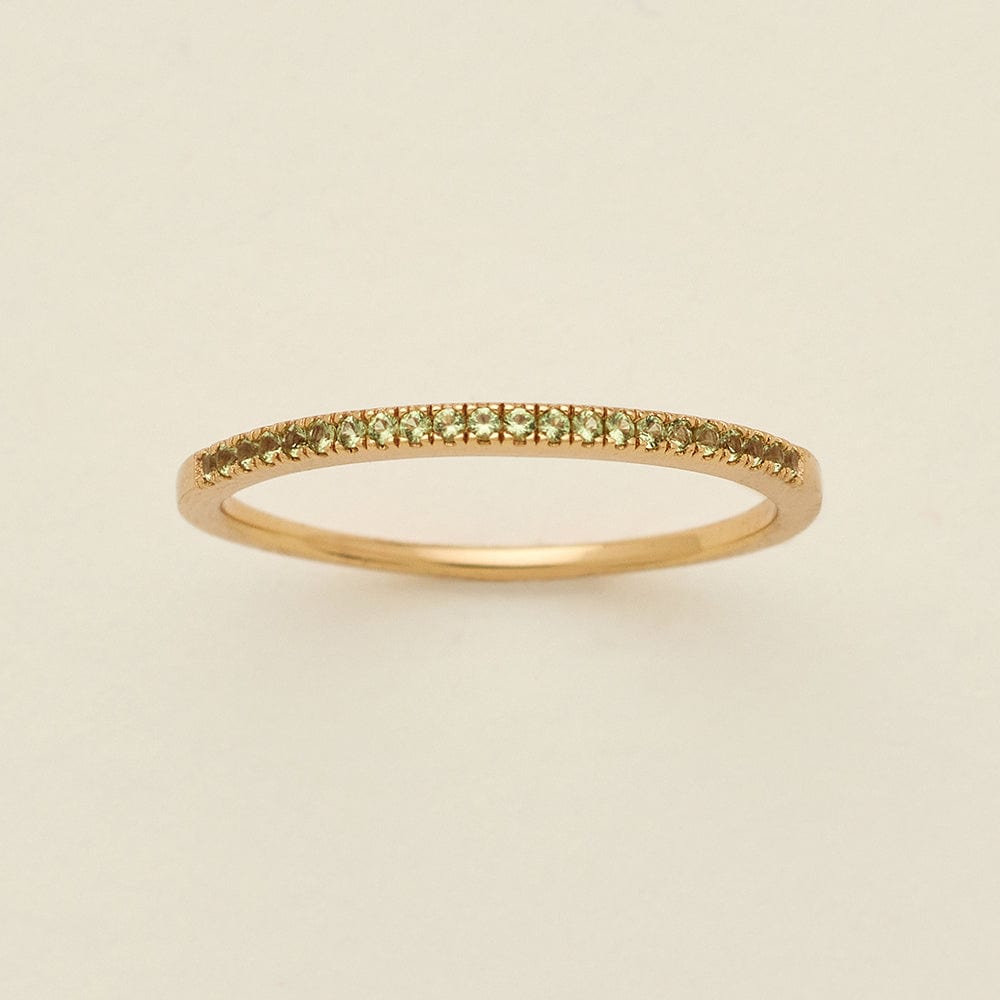 A gold ring featuring a single row of small round-cut light green gemstones, photographed on a clean, solid, warm off-white background.