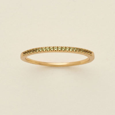 A gold ring featuring a single row of small round-cut light green gemstones, photographed on a clean, solid, warm off-white background.
