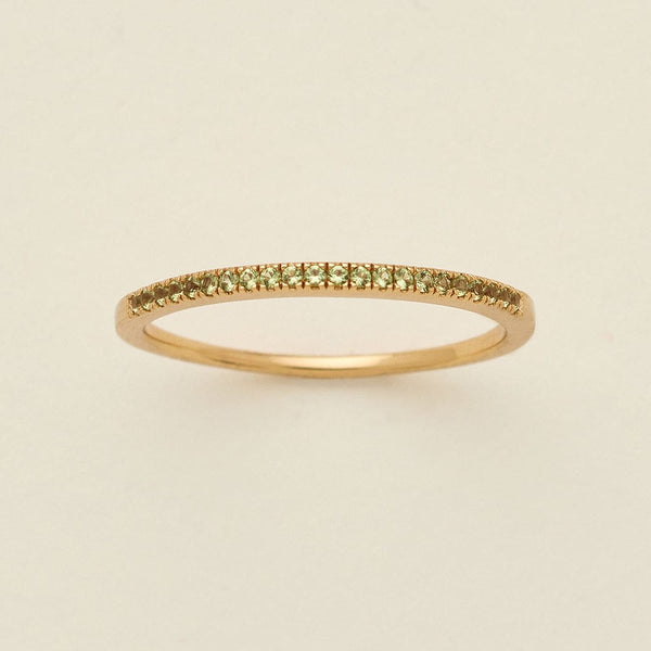 A gold ring featuring a single row of small round-cut light green gemstones, photographed on a clean, solid, warm off-white background.