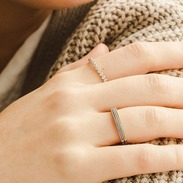 A close-up of a hand resting on a light brown thick knit sweater, showcasing a gold eternity ring made up of small round clear bezel-set stones and a stack of two thin gold rings, each with a single row of small gemstones in blue and light green.