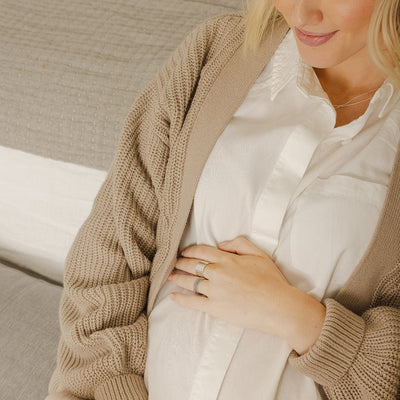 A woman in a light beige cardigan and white button-up shirt, resting her hand across her stomach and showcasing a thick polished gold cigar band ring and multiple thin stacked rings, each with a different color of gemstone in a single row across the front.