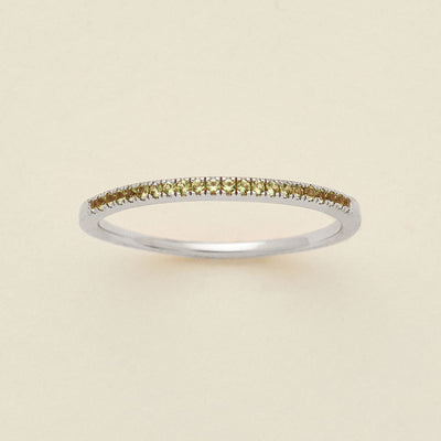 A silver ring featuring a single row of small round-cut light green gemstones, photographed on a clean, solid, warm off-white background.