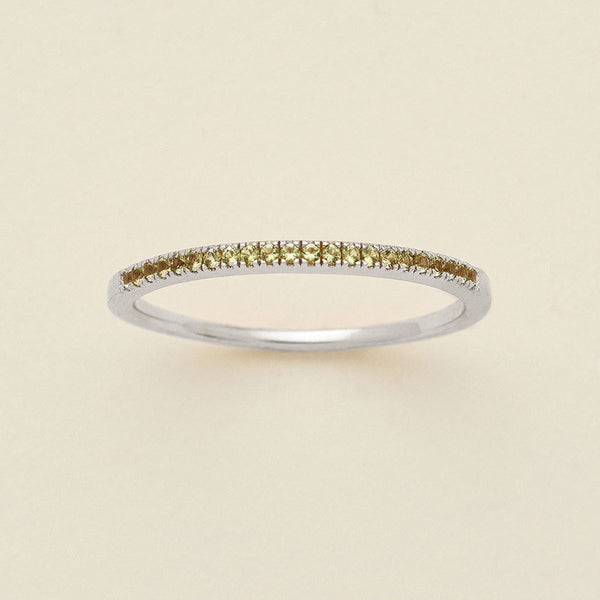 A silver ring featuring a single row of small round-cut light green gemstones, photographed on a clean, solid, warm off-white background.