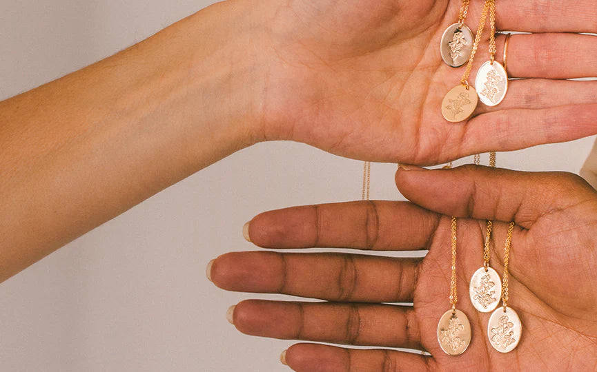 Two hands held one on top of the other, holding multiple gold pendant chain necklaces with engraved oval charms, showing a flower stem with different numbers of flowers.