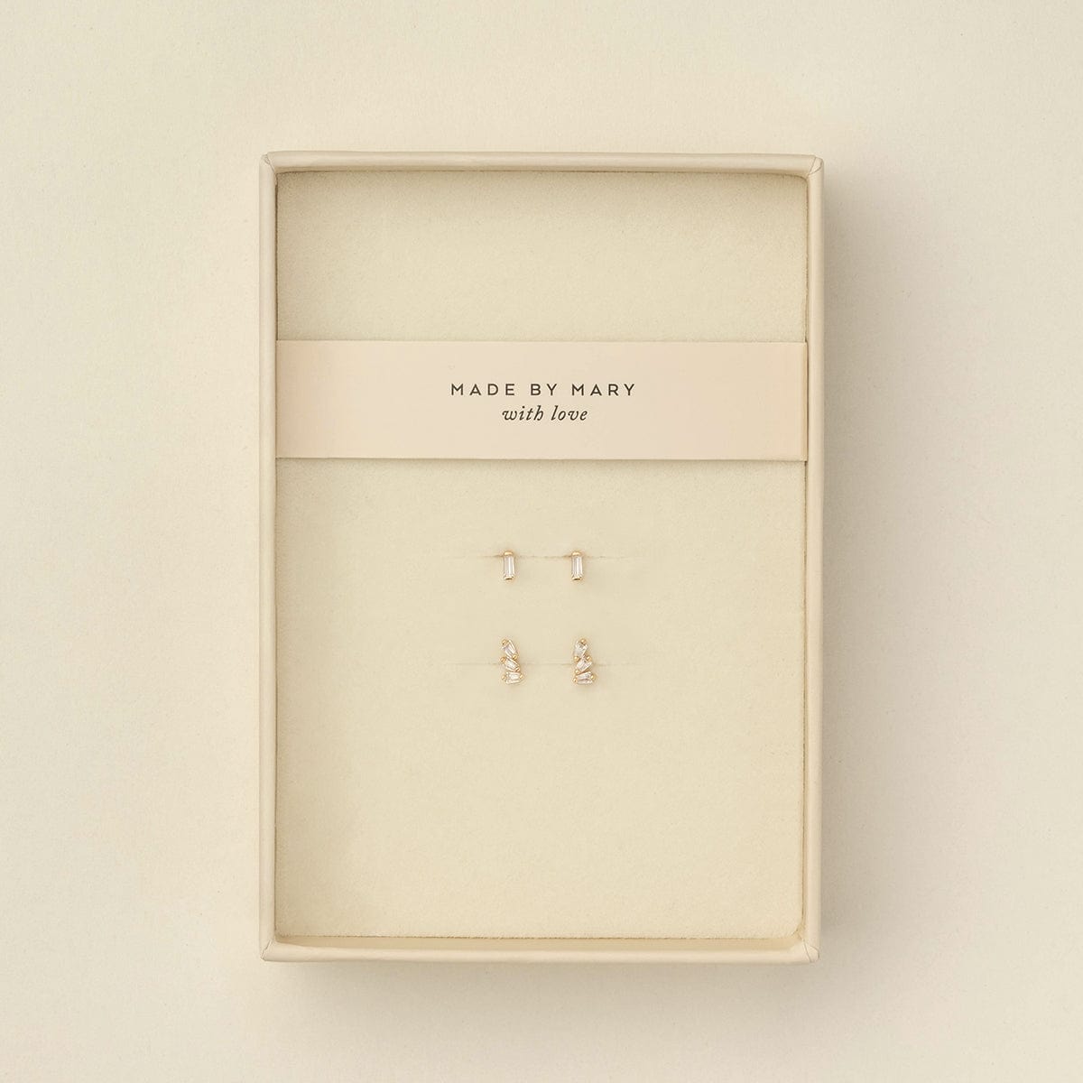 Two pairs of small gold stud earrings with baguette clear gemstone accents, displayed on a cream-colored jewelry card inside a cream-colored presentation box. The card is printed with the brand name 'MADE BY MARY with love'.
