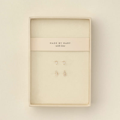 Two pairs of small gold stud earrings with baguette clear gemstone accents, displayed on a cream-colored jewelry card inside a cream-colored presentation box. The card is printed with the brand name 'MADE BY MARY with love'.