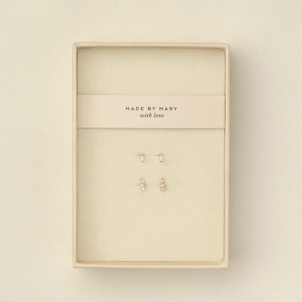 Two pairs of small gold stud earrings with baguette clear gemstone accents, displayed on a cream-colored jewelry card inside a cream-colored presentation box. The card is printed with the brand name 'MADE BY MARY with love'.