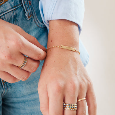A person in a light blue top and blue denim jeans wearing multiple gold rings with different blue hued stones and a gold chain bracelet with a horizontal bar.