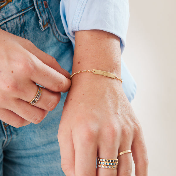 A person in a light blue top and blue denim jeans wearing multiple gold rings with different blue hued stones and a gold chain bracelet with a horizontal bar.