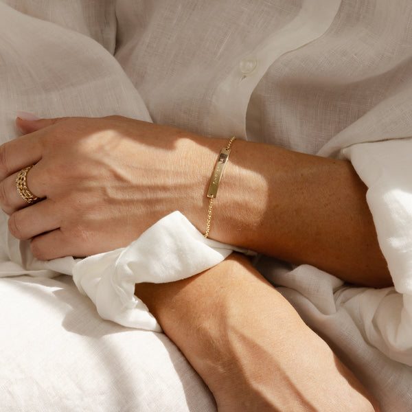 A person in a white linen button up top with their hands crossed in their lap, wearing a gold chain bracelet with an engraved horizontal bar.