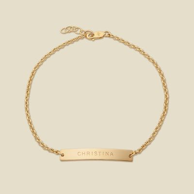 A gold chain bracelet with a horizontal bar engraved with the name 'CHRISTINA', shown on a solid light beige background.