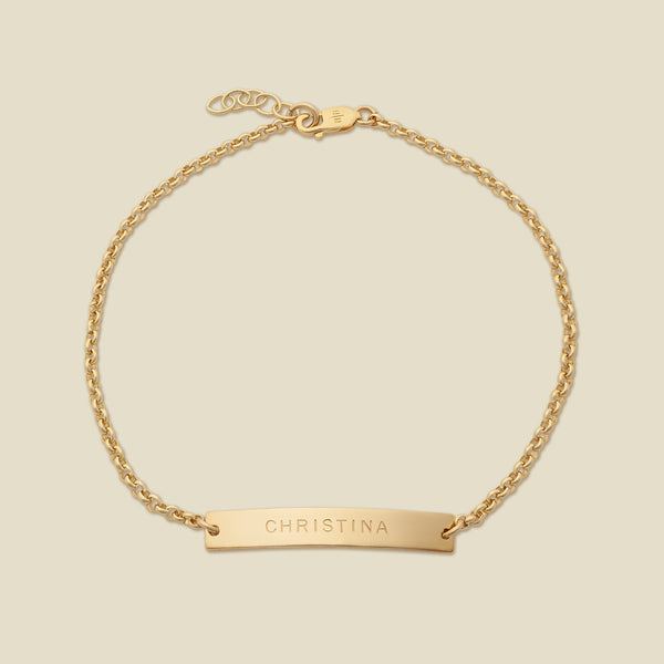 A gold chain bracelet with a horizontal bar engraved with the name 'CHRISTINA', shown on a solid light beige background.