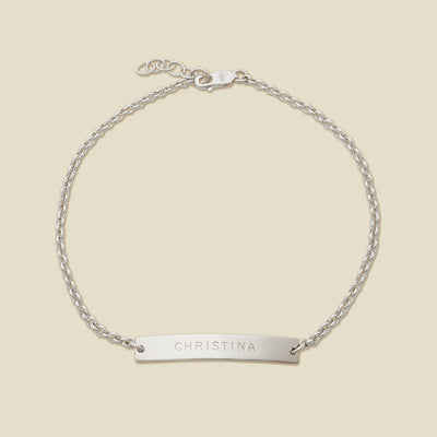 A silver chain bracelet with a horizontal bar engraved with the name 'CHRISTINA', shown on a solid light beige surface.