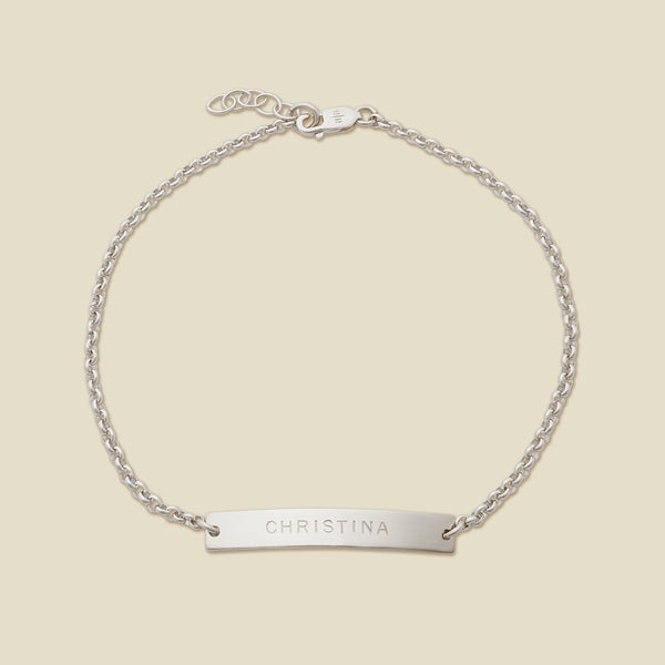 A silver chain bracelet with a horizontal bar engraved with the name 'CHRISTINA', shown on a solid light beige surface.