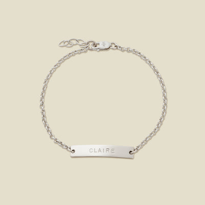 A silver chain bracelet with a horizontal bar engraved with the name 'CLAIRE' shown on a solid light beige surface.