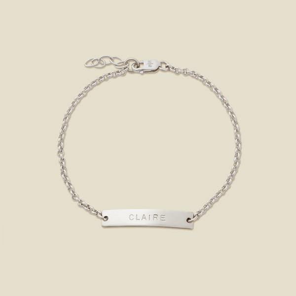 A silver chain bracelet with a horizontal bar engraved with the name 'CLAIRE' shown on a solid light beige surface.