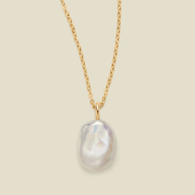 A gold chain necklace featuring a large, irregular baroque pearl pendant with a soft pearlescent luster, set against a solid light beige background.