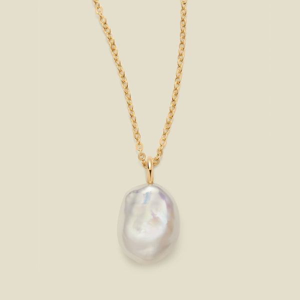 A gold chain necklace featuring a large, irregular baroque pearl pendant with a soft pearlescent luster, set against a solid light beige background.