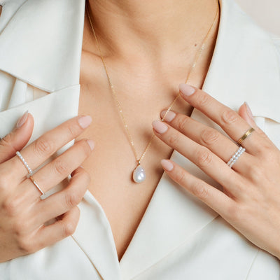A close-up of a person's neck wearing a gold pendant chain necklace with a large organic pearl charm, paired with multiple pearl strand rings and gold thin band rings.