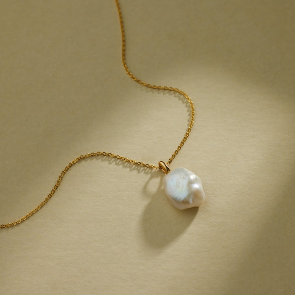 A gold pendant chain necklace with a large organic pearl charm, laying against a textured beige background with natural shadows.