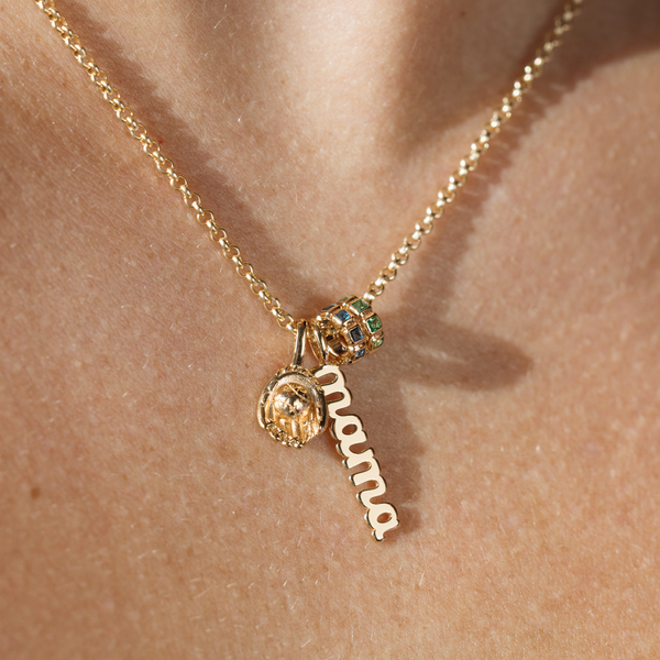 A close-up image of a person wearing a gold chain necklace with a gold baseball mitt charm, a gold script font charm with the word 'mama' hanging vertically, and two ring charms comprised of square, bezel-set stones. One is blue-green, and the other is emerald green.