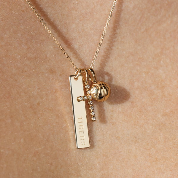 A close-up image of a person wearing a gold pendant chain with a vertical bar charm engraved with the word 'TIGERS', a gold cross charm with clear gemstones, and a gold basketball charm.