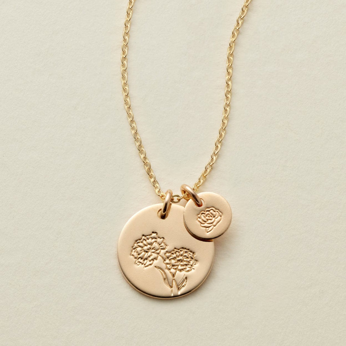 A gold chain necklace with two round flat discs, one larger and one smaller, each engraved with a different floral design and shown on a solid light beige background.