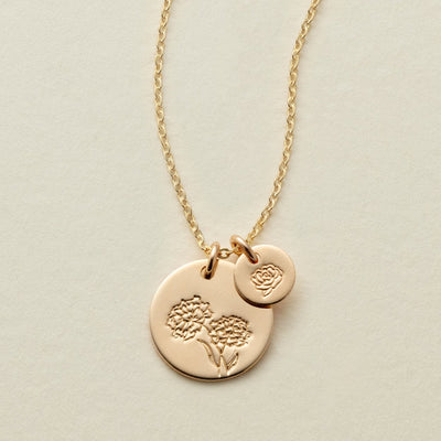 A gold chain necklace with two round flat discs, one larger and one smaller, each engraved with a different floral design and shown on a solid light beige background.