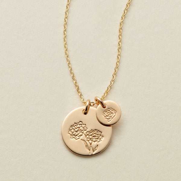 A gold chain necklace with two round flat discs, one larger and one smaller, each engraved with a different floral design and shown on a solid light beige background.