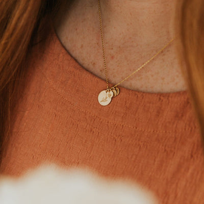 A close-up of a woman in a burnt orange top wearing a delicate gold pendant chain with one large flat round disc and three smaller flat round discs. Each disc is engraved with a different floral pattern.