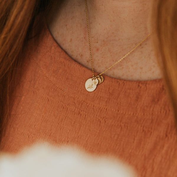 A close-up of a woman in a burnt orange top wearing a delicate gold pendant chain with one large flat round disc and three smaller flat round discs. Each disc is engraved with a different floral pattern.