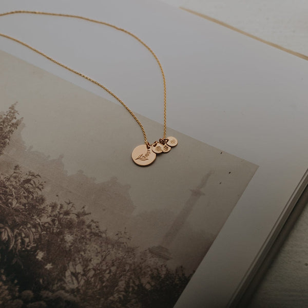 An open page of a black and white picture book showing a landscape, with a delicate gold pendant chain necklace laying across it. The necklace has four round flat discs, one larger and three smaller, each with a different floral design.