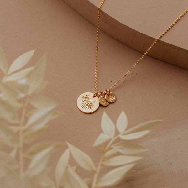 A gold chain necklace with three round flat discs, one larger and two smaller, each engraved with a different floral design and shown on a beige backdrop with foliage blurred in the foreground.