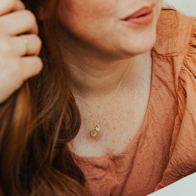 A close-up of a woman in a burnt orange top wearing a delicate gold pendant chain necklace with two round flat discs, one smaller and one larger. Each disc features an engraved floral design.