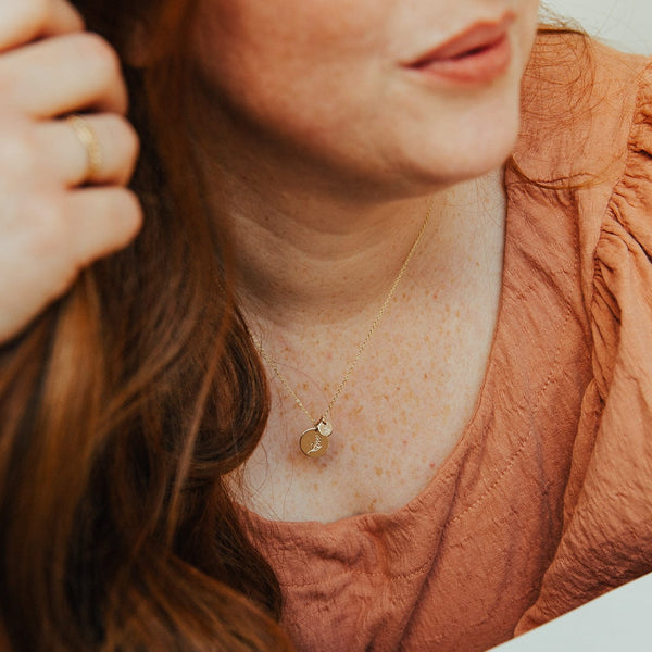 A close-up of a woman in a burnt orange top wearing a delicate gold pendant chain necklace with two round flat discs, one smaller and one larger. Each disc features an engraved floral design.