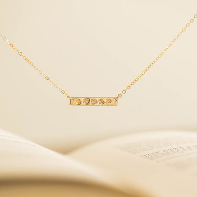 A delicate gold chain necklace with a stationary horizontal bar, engraved with different birth flower designs and shown hanging in front of an off-white background.