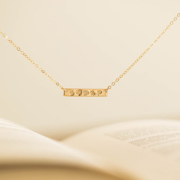 A delicate gold chain necklace with a stationary horizontal bar, engraved with different birth flower designs and shown hanging in front of an off-white background.