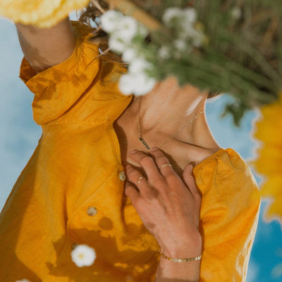 A woman's reflection in a mirror wearing a golden yellow top with a gold bar necklace engraved with flowers.