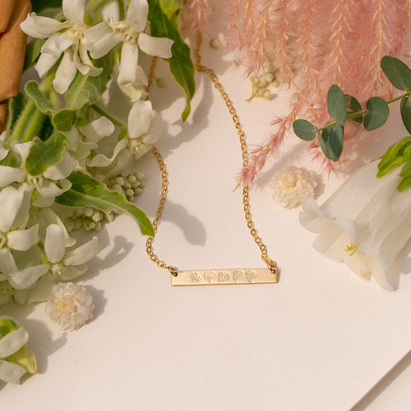 A delicate gold chain necklace with a stationary horizontal bar engraved with birth flower designs, laying on a solid white surface with white, green, and pink flowers and foliage surrounding it.