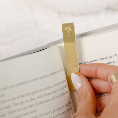 A close-up of a person putting a gold bookmark into the page of a book. The bookmark features an engraved floral design and initials on the top.