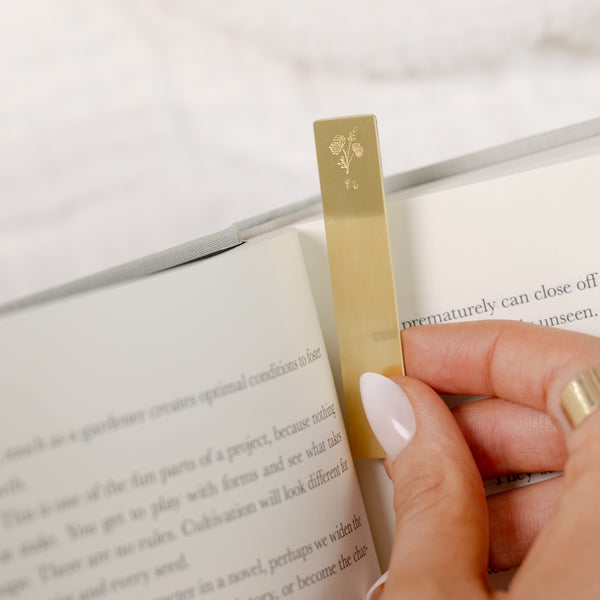 A close-up of a person putting a gold bookmark into the page of a book. The bookmark features an engraved floral design and initials on the top.