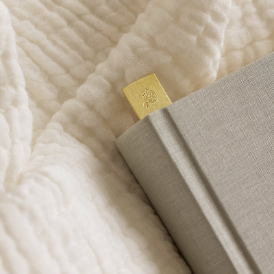 A close-up of the corner of a book laying on a white blanket, showcasing the gold bookmark with an engraved peony.