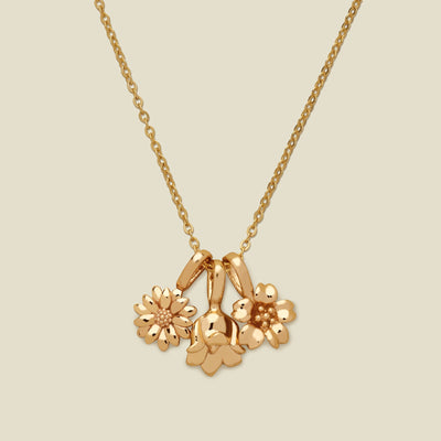 A gold pendant necklace with three small flower charms, shown on a solid light beige background.
