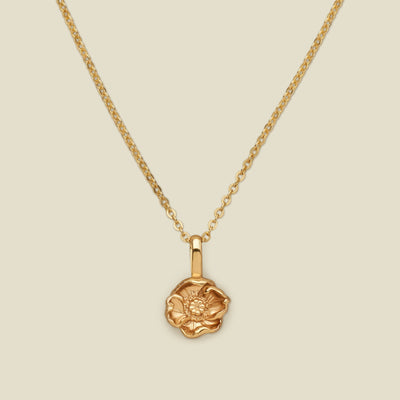 A gold pendant necklace with a small poppy pendant charm, shown on a solid light beige background.