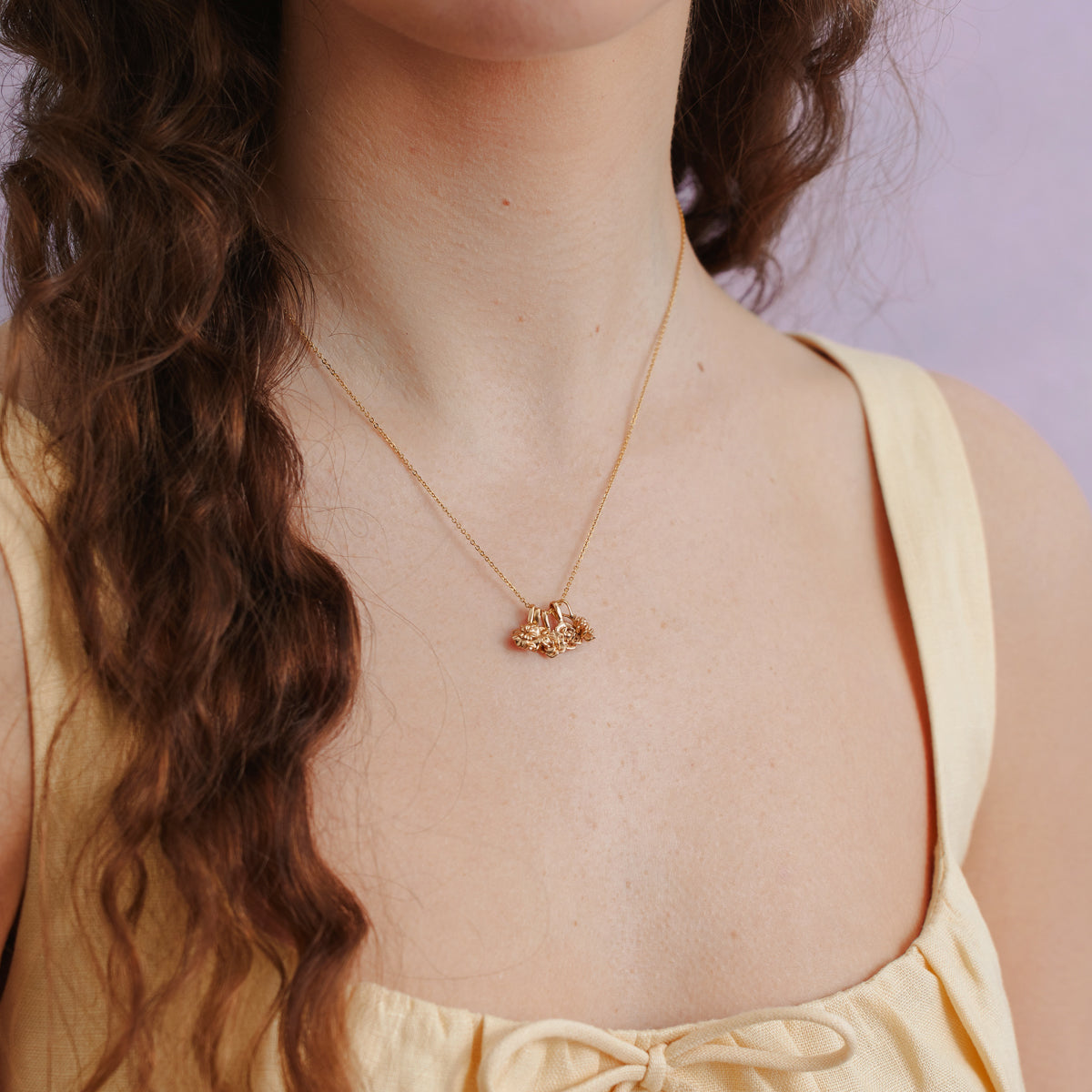 A close-up of a woman in a pastel yellow top wearing a delicate gold chain necklace with three gold flower charms with a light purple background.