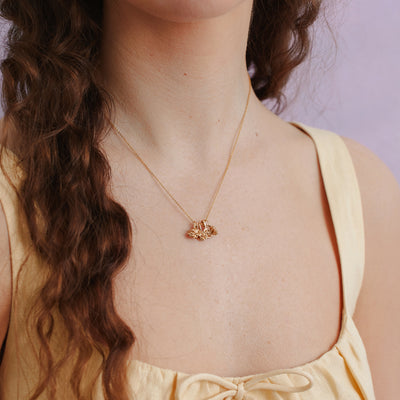A close-up of a woman in a pastel yellow top wearing a delicate gold chain necklace with three gold flower charms with a light purple background.