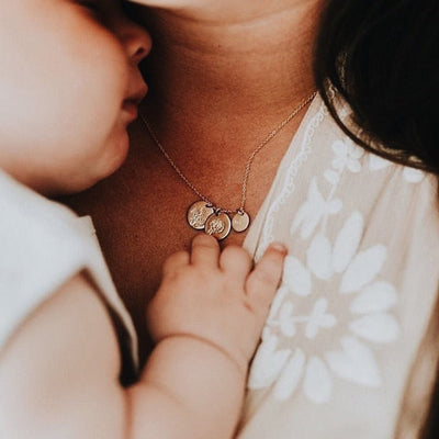 A close-up of a woman with a small child on her chest, showcasing her delicate gold pendant chain necklace with three round flat discs. Each disc features a different floral motif engraving.