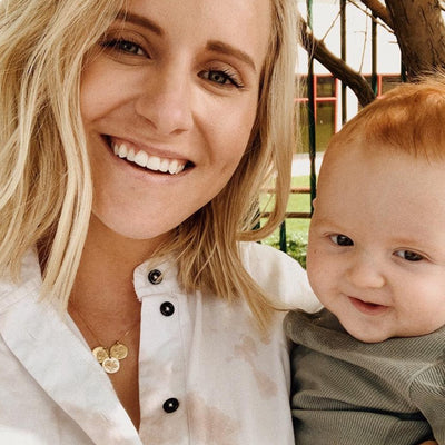 A blonde woman wearing a white button up top, holding her small child and showcasing her delicate gold pendant chain necklace with three round flat discs, each with a different floral design engraved.