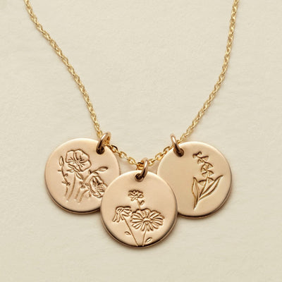 A gold chain necklace with three round engraved disc pendants featuring floral  designs, displayed on a light beige background.