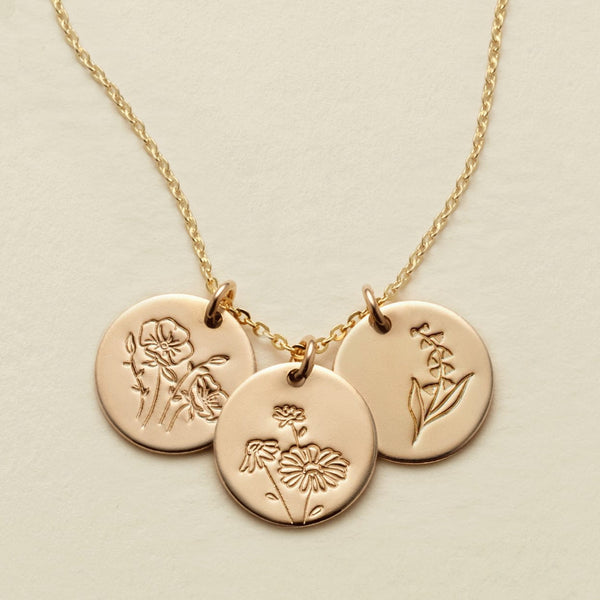 A gold chain necklace with three round engraved disc pendants featuring floral  designs, displayed on a light beige background.